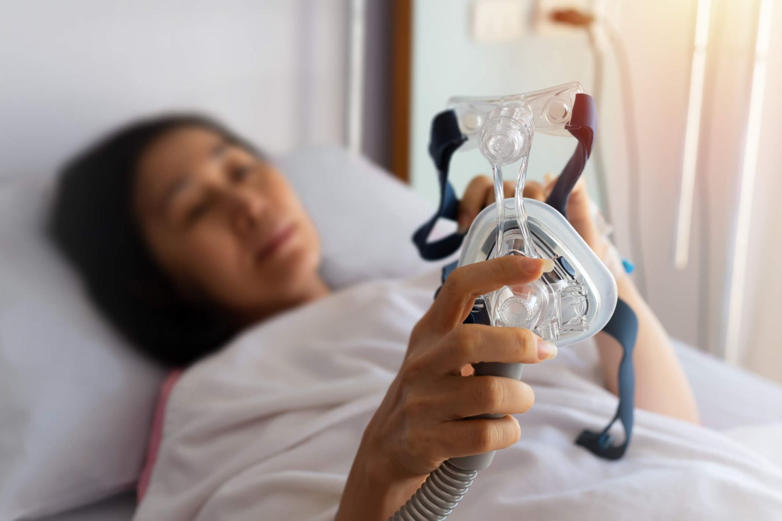 Asian woman lying in bed holding CPAP mask in hands